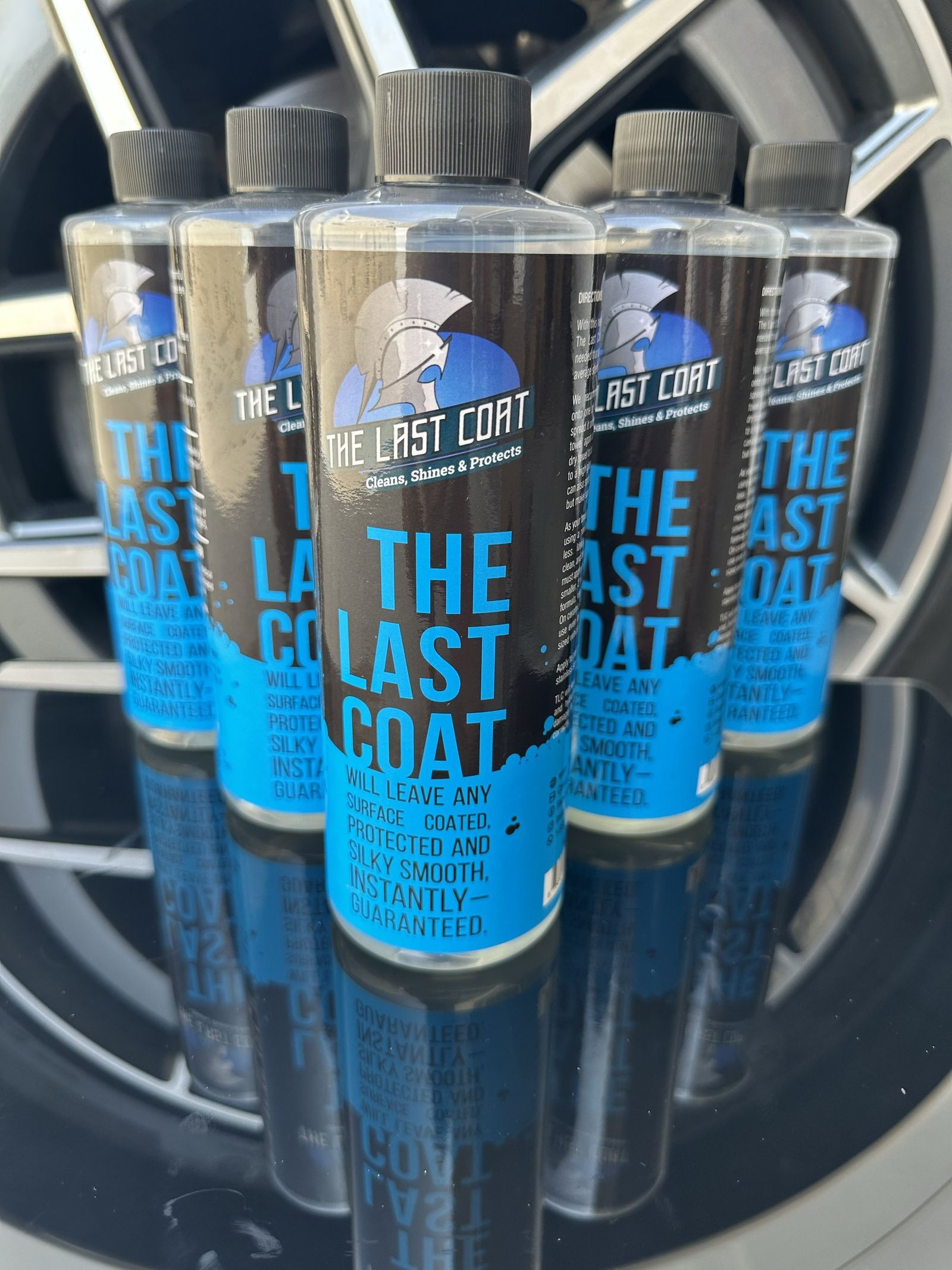 Five bottles of The Last Coat 2.0 on a black reflective surface in front of a car wheel